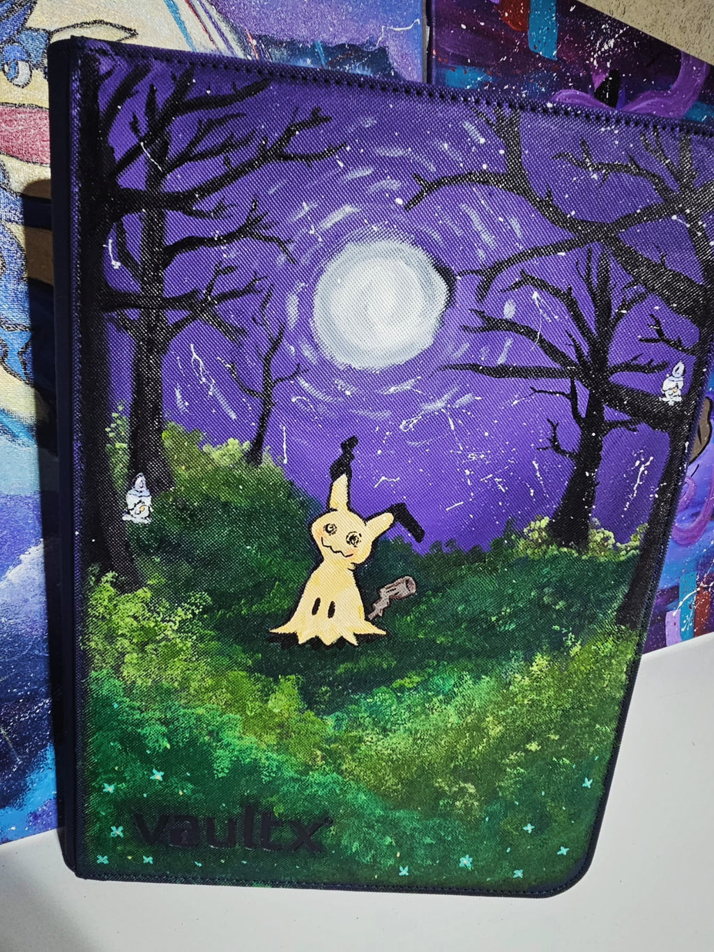 Angled front view of custom hand-painted Mimikyu VaultX binder — yellow Mimikyu standing in moonlit forest clearing under full moon on deep purple starry night sky, litwick hiding in a tree, freehand acrylic artwork by 5/14 Apparel