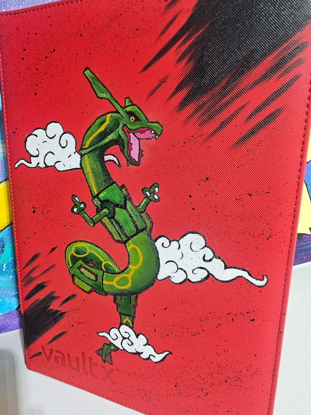 Angled front view of custom hand-painted Rayquaza VaultX binder — green Rayquaza soaring through white clouds on bold red background, freehand acrylic artwork by 5/14 Apparel