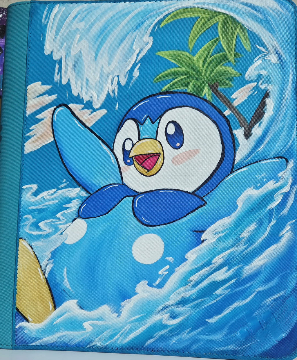 Close view of 5/14 Apparel custom hand-painted Piplup binder — Piplup riding a wave with crashing water and tropical island, one-of-one acrylic art on teal 9-pocket top loader binder