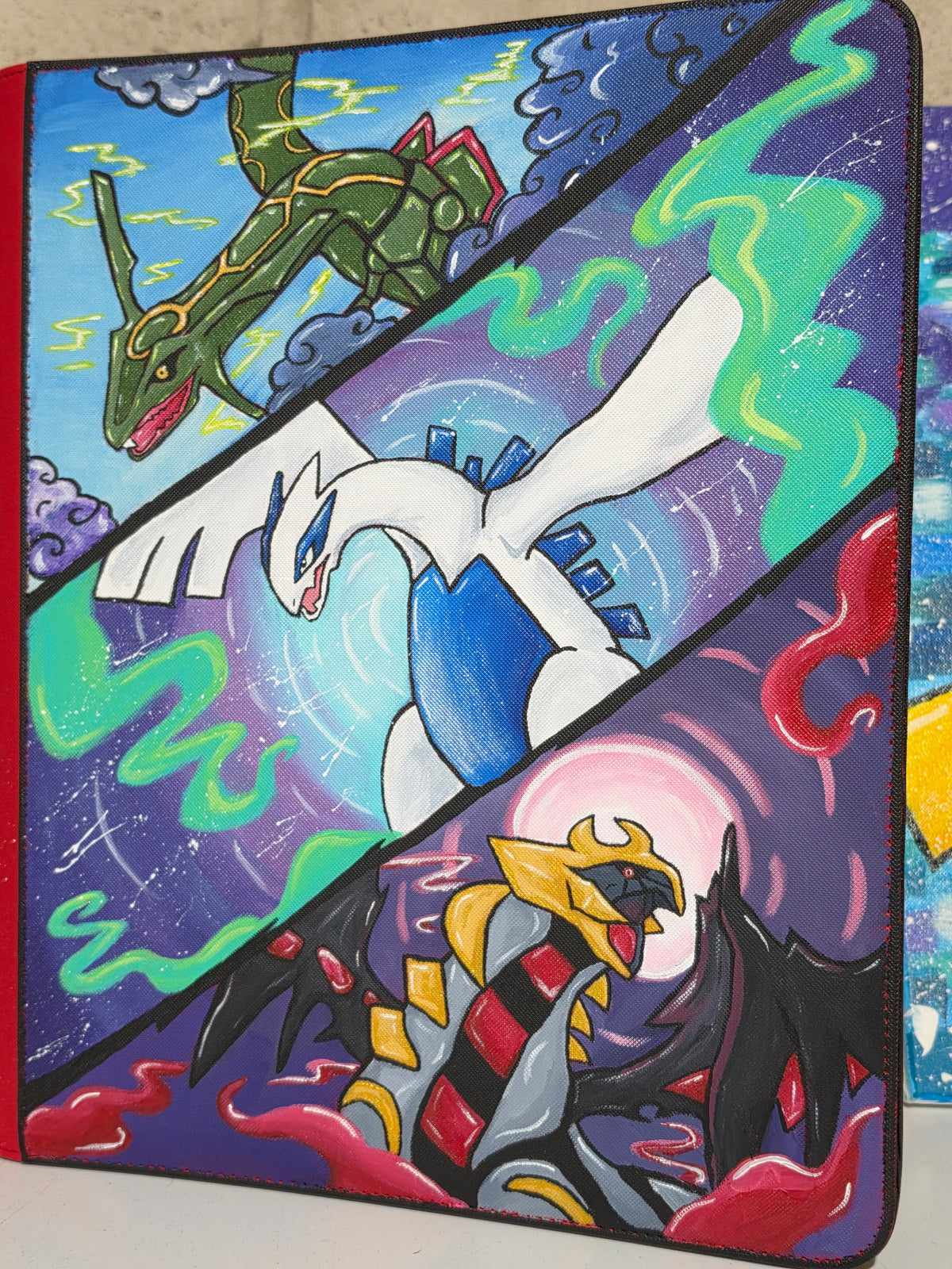 Close view of 5/14 Apparel custom hand-painted Rayquaza Lugia and Giratina binder — detailed legendary trio panel artwork with bold colors and dynamic energy effects, one-of-one acrylic art on red 9-pocket binder