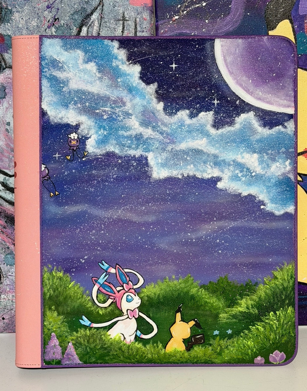Front view of 5/14 Apparel custom hand-painted Sylveon and Mimikyu binder — Sylveon and Mimikyu sitting in moonlit green meadow under deep purple starry sky with large crescent moon, one-of-one acrylic art on pink 9-pocket binder