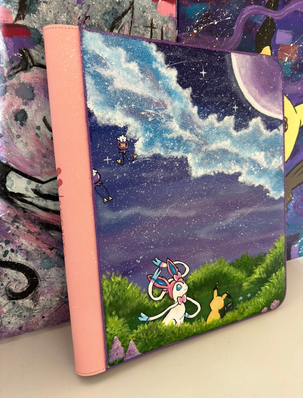Side view of 5/14 Apparel custom hand-painted Sylveon and Mimikyu binder — Sylveon and Mimikyu in starlit meadow with Drifloon floating above under purple night sky and crescent moon, one-of-one acrylic art on pink 9-pocket binder