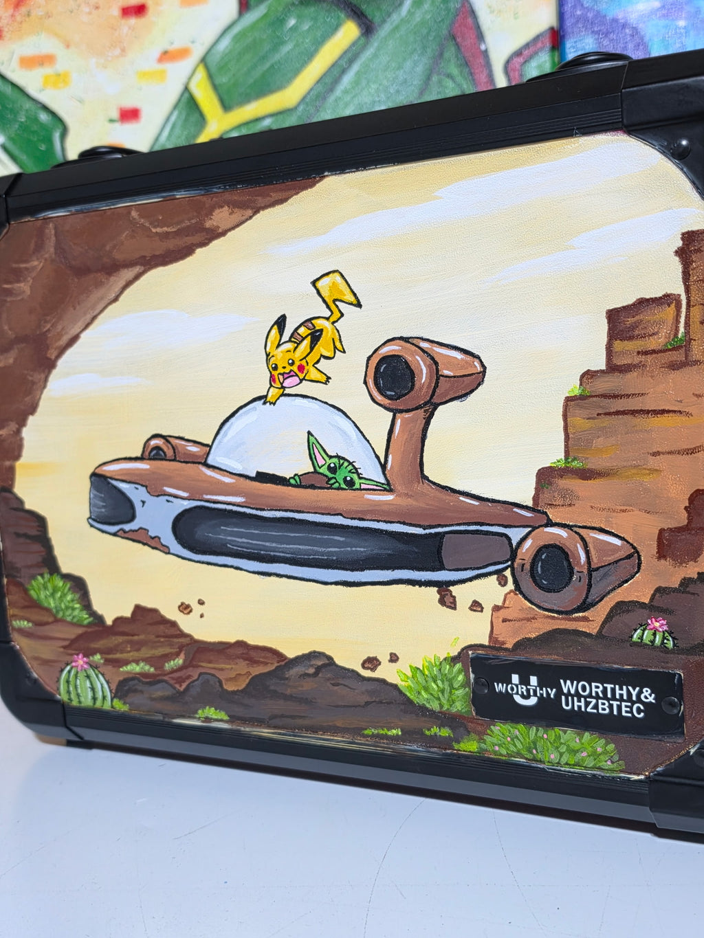 Close-up angle of hand-painted Pikachu & Grogu Razor Crest slab case — freehand acrylic art detail showing canyon scene, Worthy & Uhzbtec collab badge, black case


